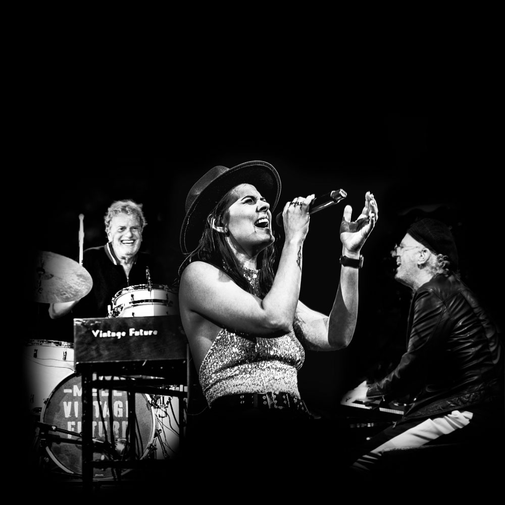Zwart witte foto van de band met twee manen achter een drumstel en piano en een zangeres