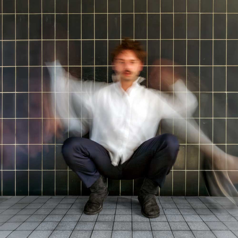 Voorstellingsbeeld van Donny Ronny: 'Man in wazige beweging zittend in lotushouding achter een glazen raster'.
