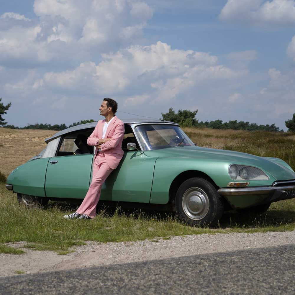 Voorstellingsbeeld van Gerard Alderliefste: man in roze pak leunend tegen klassieke groene auto, met landschap op de achtergrond.