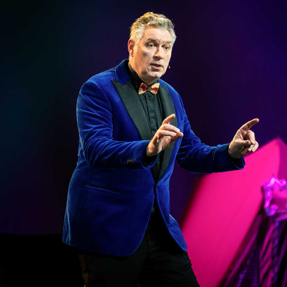 Voorstellingsbeeld van Sjaak Bral: 'Man in blauw pak en rode vlinderdas spreekt op een podium met microfoonheadset.