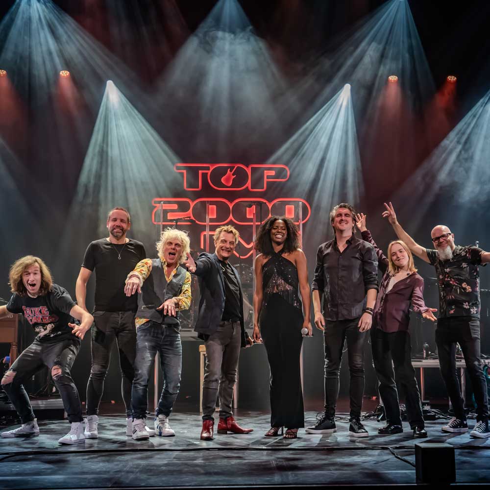 Voorstellingsbeeld van Top 2000 live: 'Bandleden lachen en gebaren naar de camera voor een TOP 2000 logo op het podium.