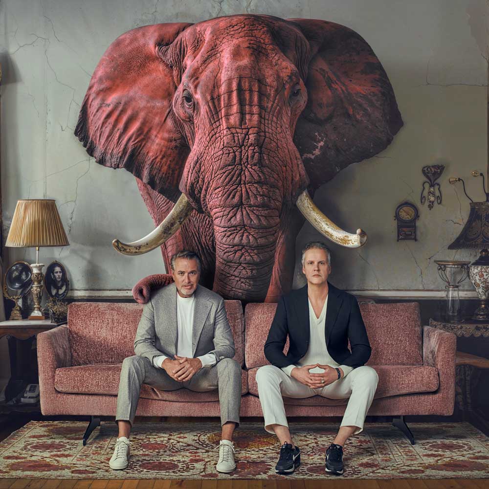 Voorstellingsbeeld van Veldhuis en Kemper: 'Twee mannen zittend op een bank onder een gigantische olifantenhoofd aan de muur in een vintage kamer.