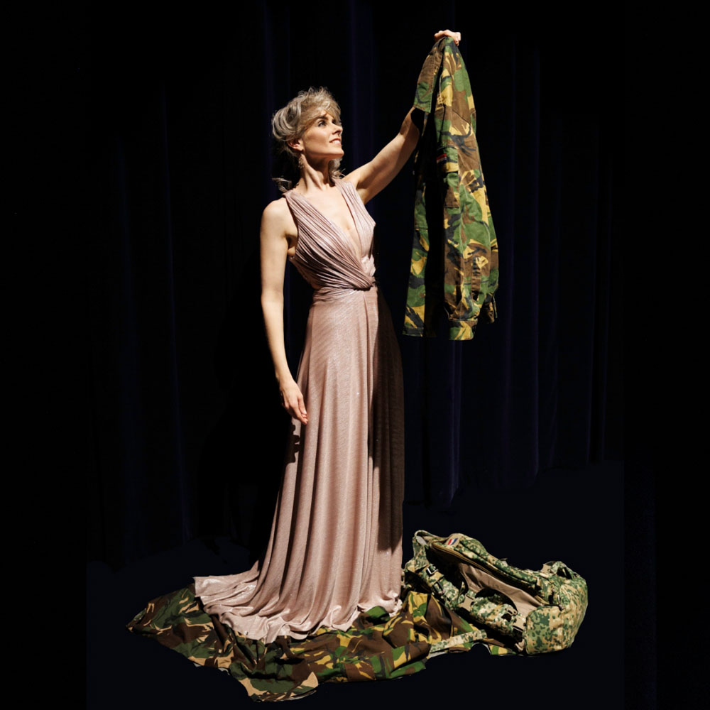 Vrouw in avondjurk houdt camouflagejas omhoog op donker podium.