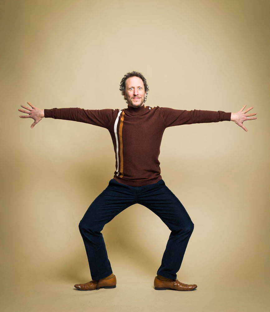 Micha Wertheim staat wijdbeens met gestrekte armen in een beige studio.