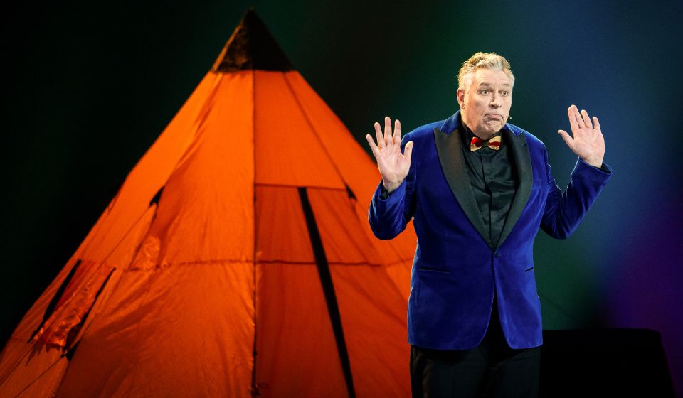 Voorstellingsbeeld van een persoon in een blauw jasje die een presentatie geeft naast een grote, verlichte oranje tipi-tent.