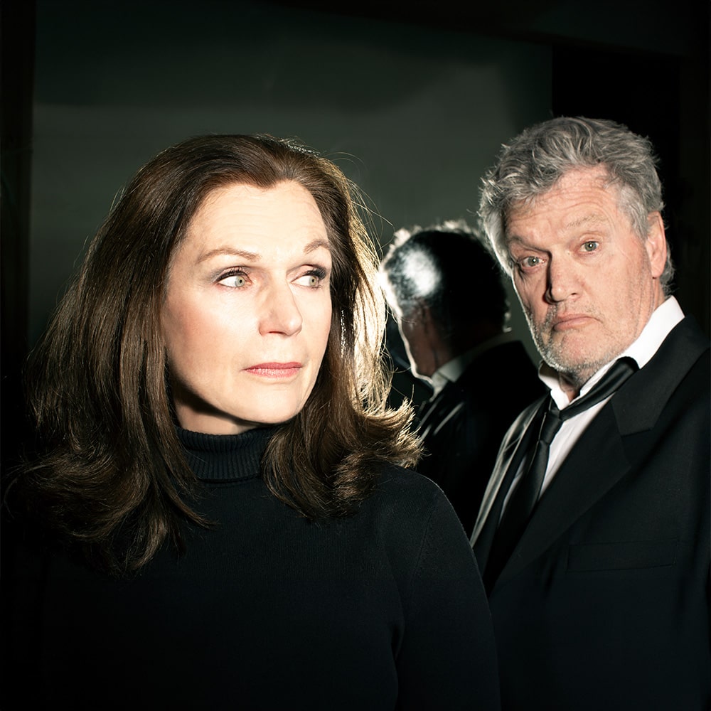 Voorstellingsbeeld van Doek; twee personen, gekleed in zwarte kledij, die bezorgd kijken tegen een verlichte achtergrond.