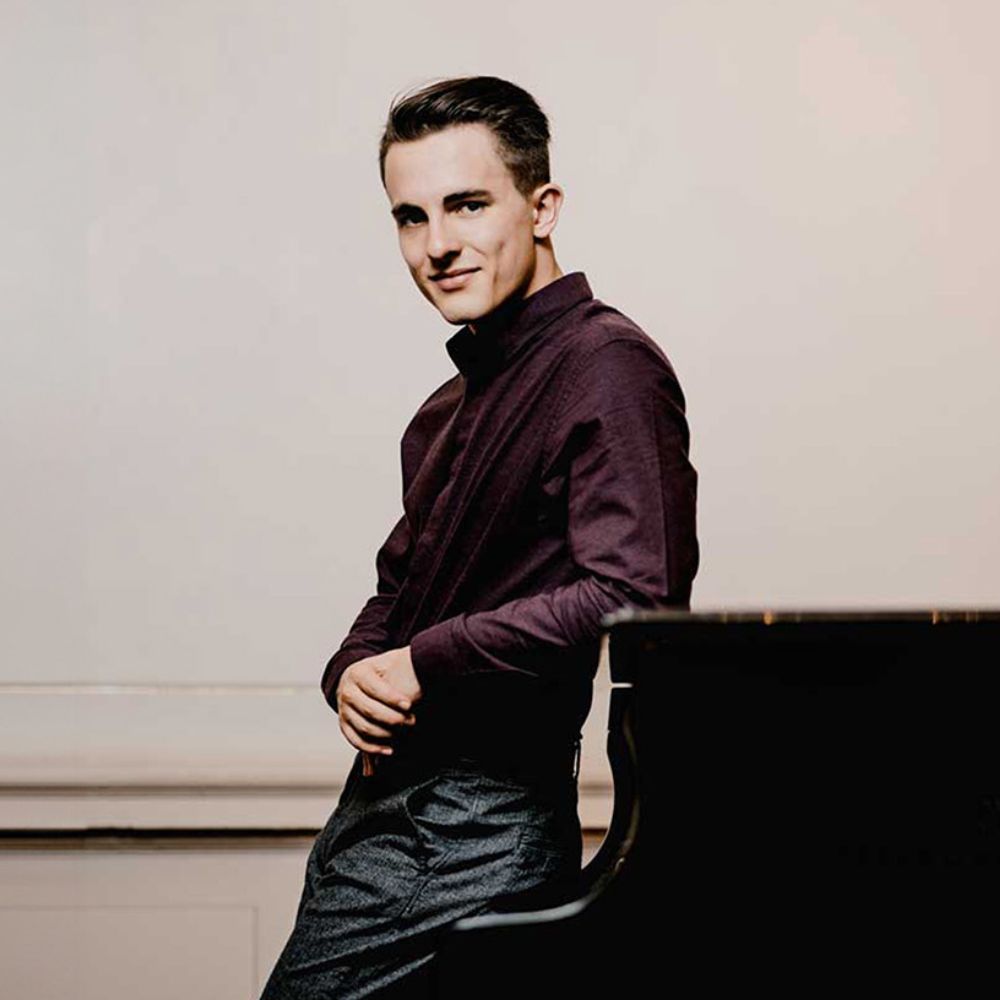 Voorstellingsbeeld van Florian Verweij: een jonge man in een donkerpaarse blouse die glimlachend over zijn schouder kijkt in een kamer met een piano op de achtergrond.