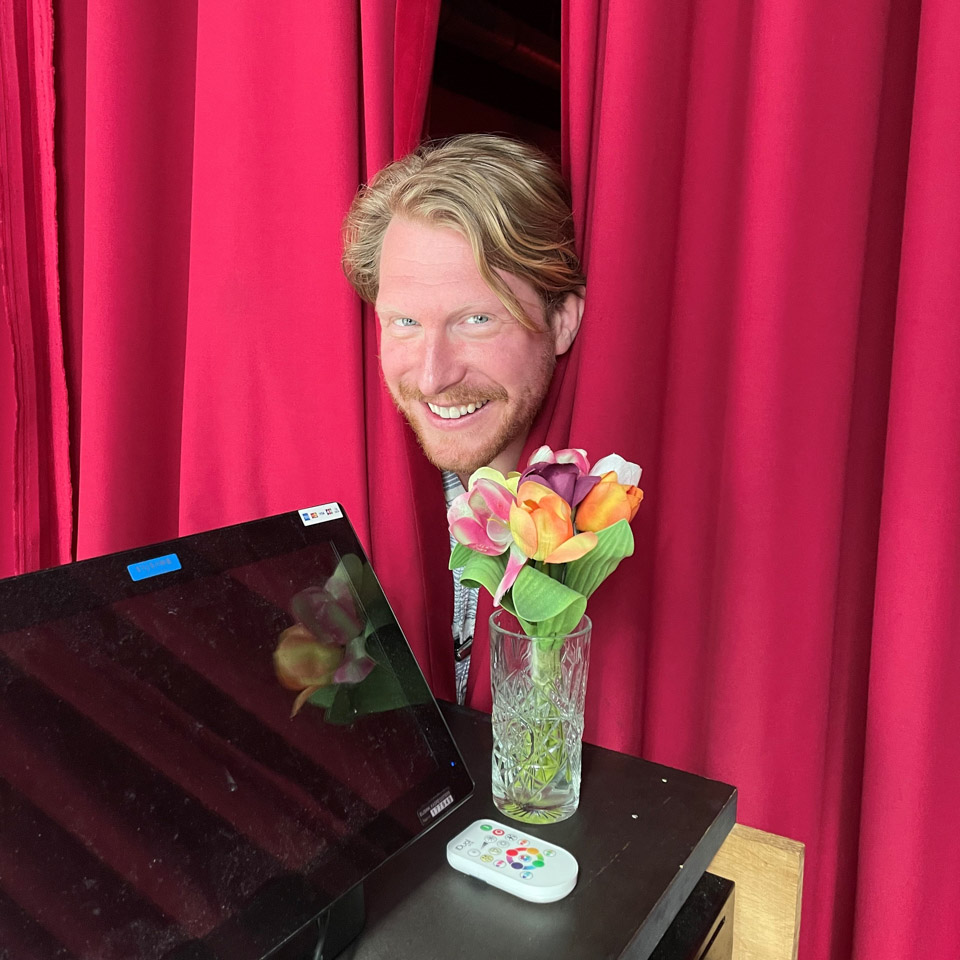 Kor Hoebe, glimlachend achter een rood gordijn, met bloemen en een afstandsbediening op tafel. Promotiefoto voor zijn voorstelling 'KORDAAT'.