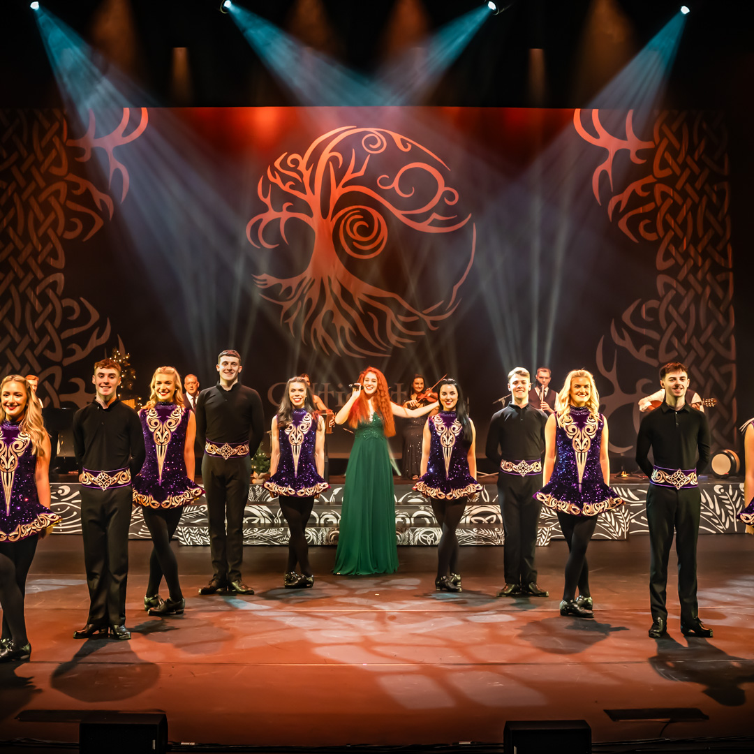 Ierse dansers in paarse kostuums en zangeres op het podium met Keltisch decor.
