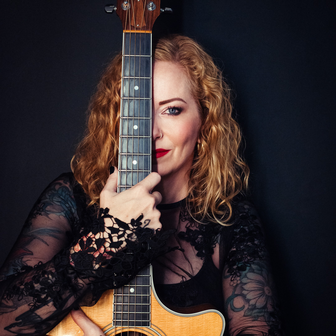 Anneke poseert met akoestische gitaar en één oog zichtbaar achter de snaren.