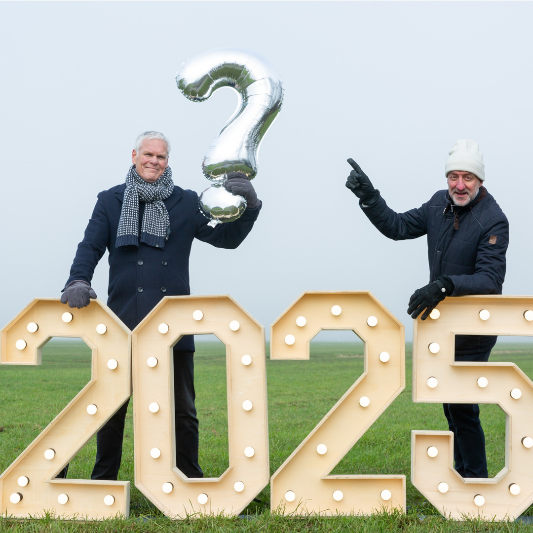 Rob Urgert en Joep van Deudekom staan bij lichtcijfers 2025, één houdt een ballon in de vorm van een vraagteken.