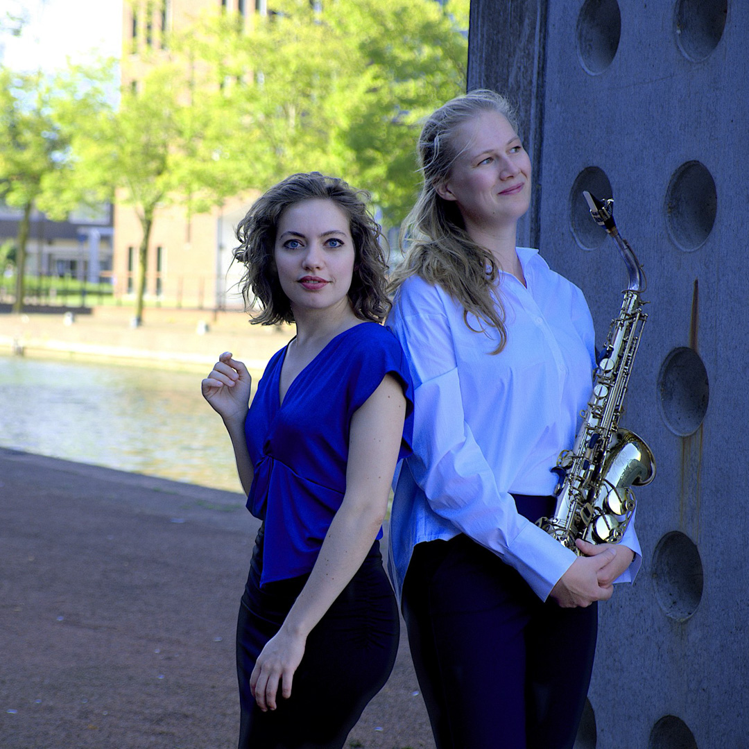 Twee muzikanten poseren buiten, één met saxofoon, de ander in felblauwe top.