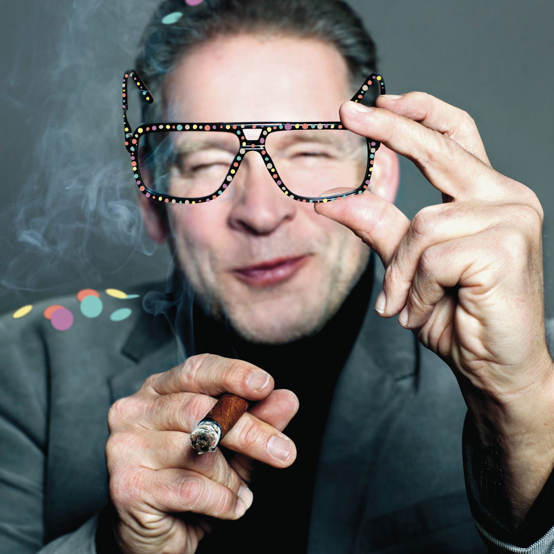 Erik van Muiswinkel rookt sigaar en houdt feestbril voor zijn gezicht.
