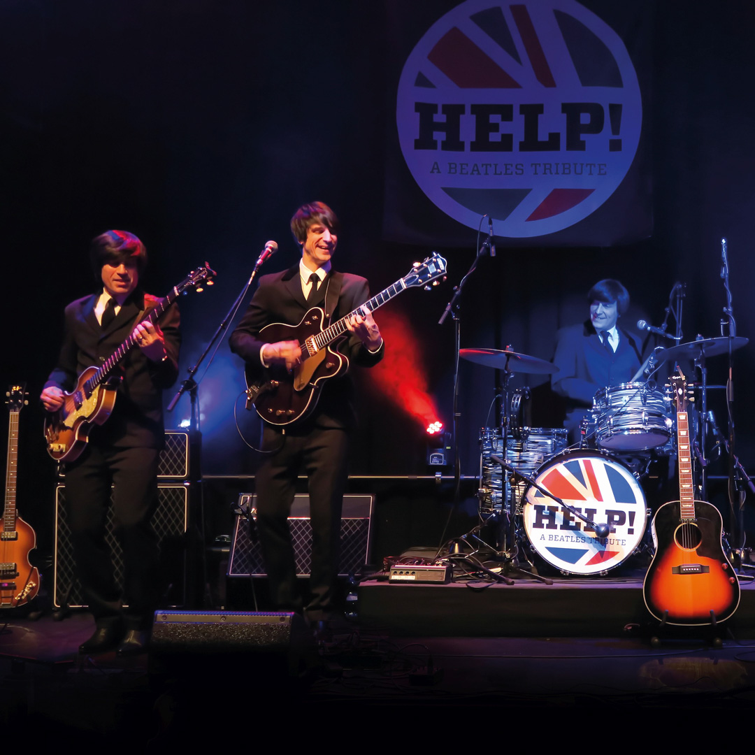 De drie gitaristen en drummer treden op onder rood en blauw licht bij HELP! – A Beatles Tribute.