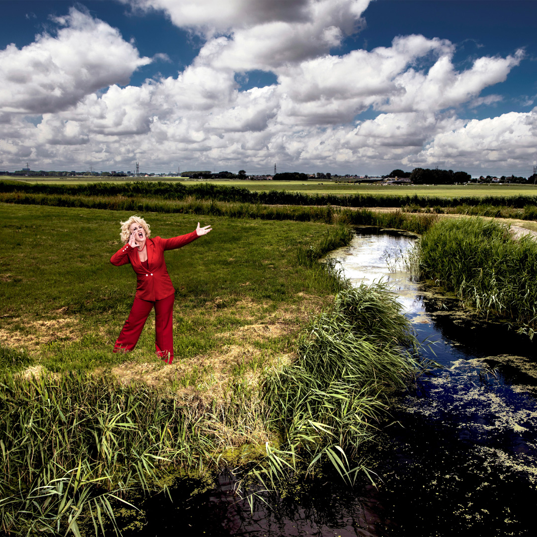 Karin Bloemen roept luid in een weids polderlandschap met sloot en wolkenlucht.