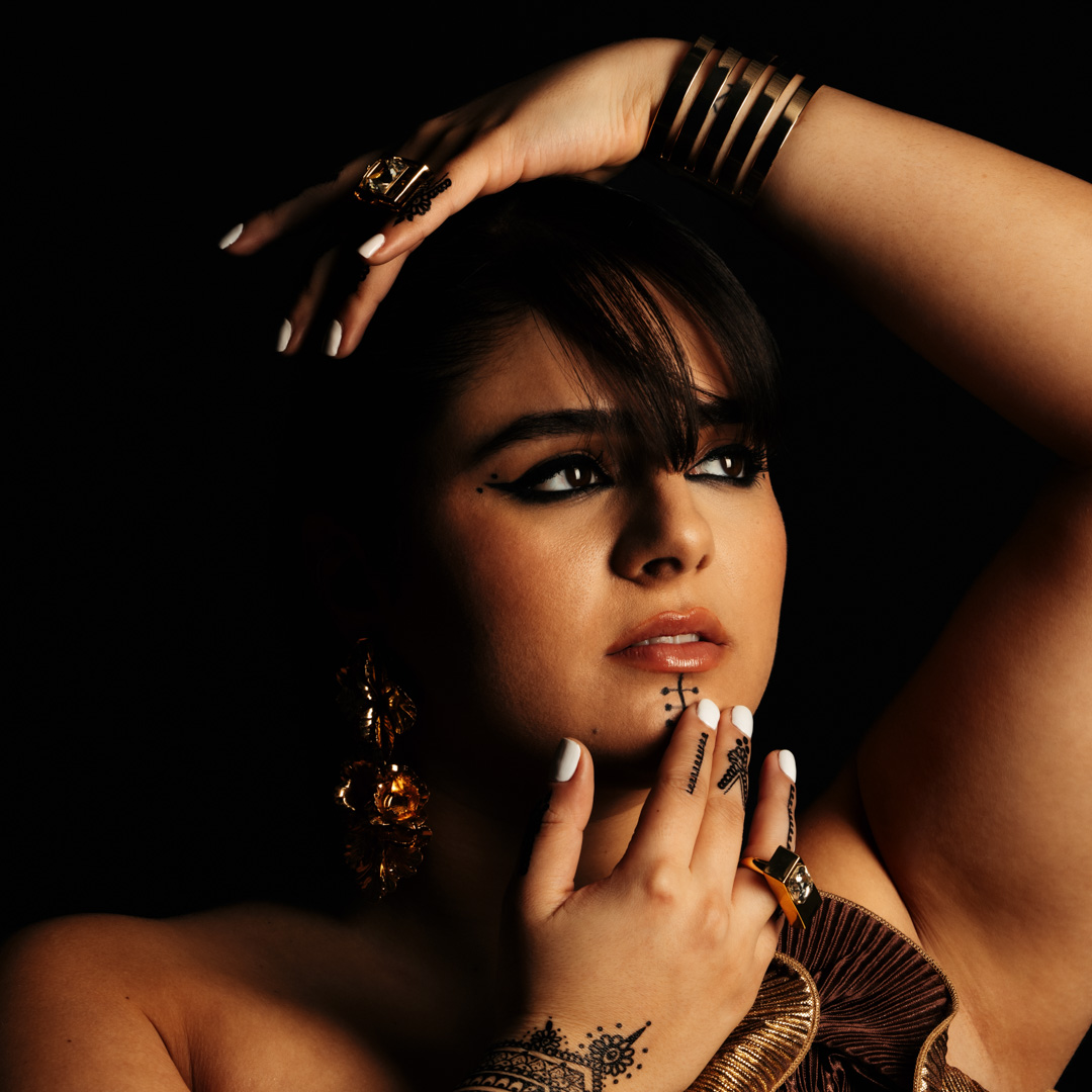 Zangeres Numidia kijkt intens omhoog met haar hand op haar kin, wit gelakte nagels en sierlijke henna op haar vingers. Ze draagt een gouden, sierlijke top.