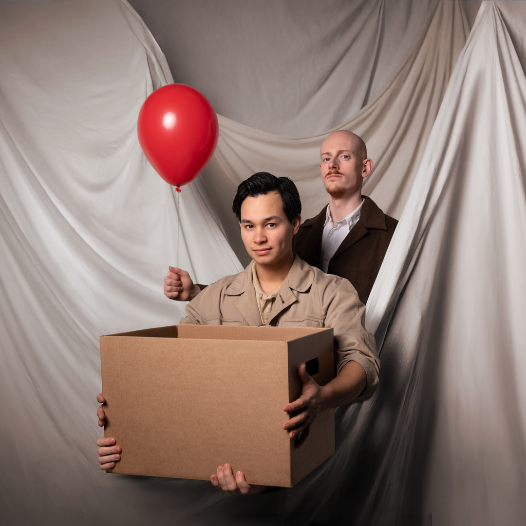 Niek heeft een rode ballon in zijn hand en Guy een kartonnen doos. De mannen poseren voor een wit doek.