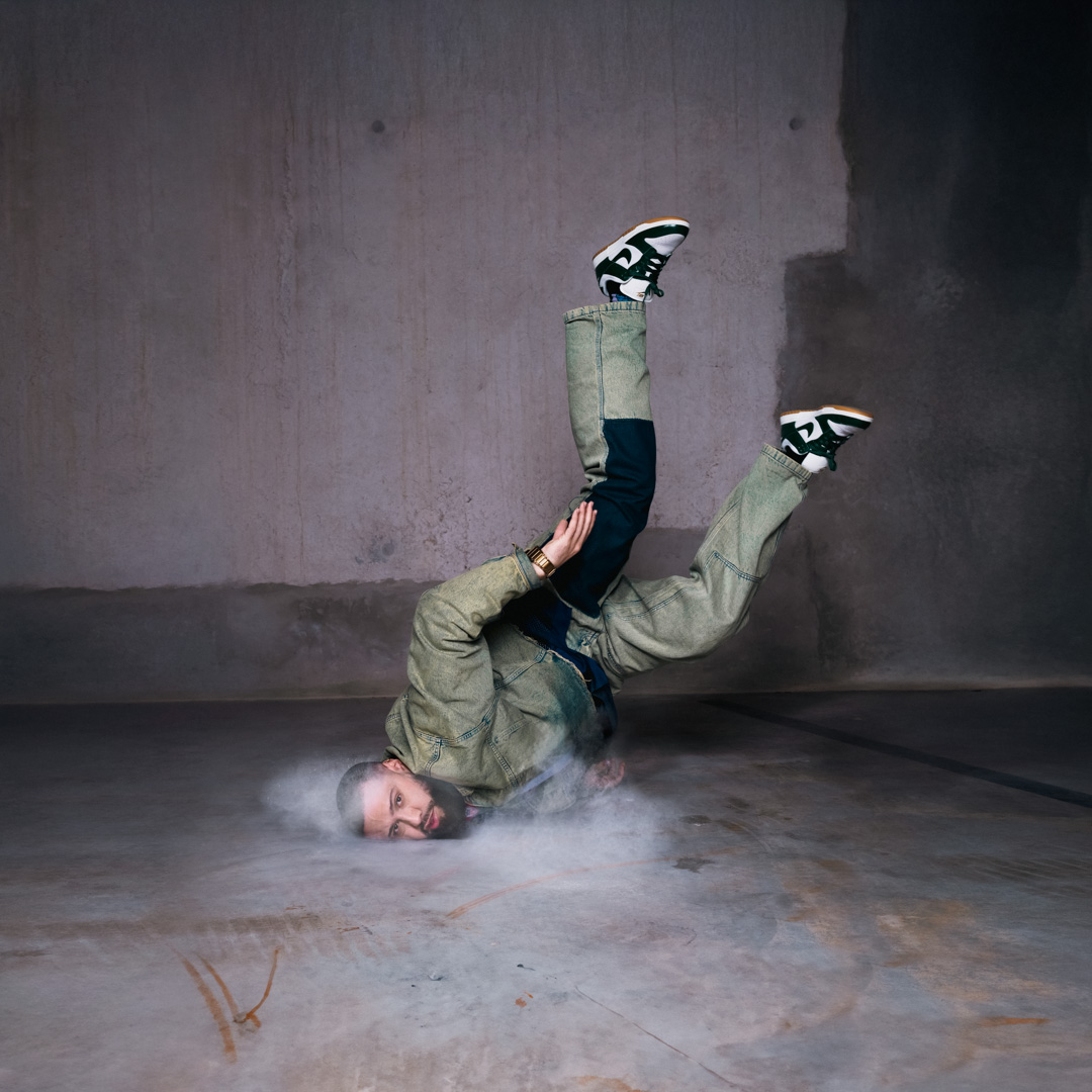 Patrick Ribeiro maakt een breakdance move met hoofd op de vloer en benen in de lucht. Hij bevindt zich in een betonnen kamer en er beweegt stof rondom zijn hoofd.
