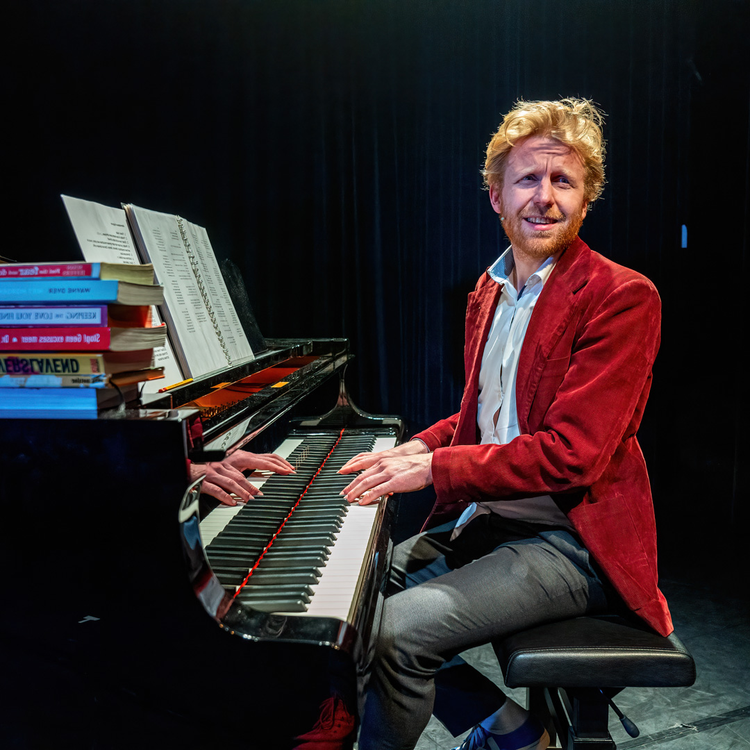 Piet van Eeghen speelt piano op een donker podium met boeken en bladmuziek.