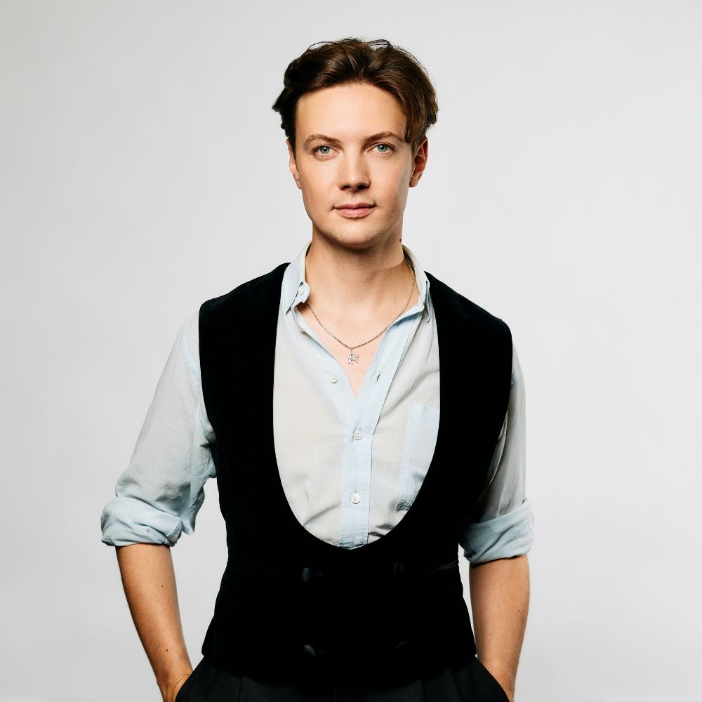 Florian Myjer kijkt recht in de camera met open overhemd en zwarte vest tegen witte achtergrond.