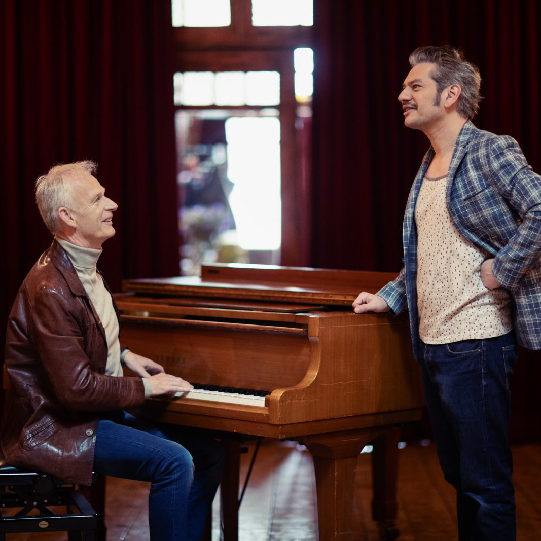 Cor Bakker speelt piano terwijl Maarten Heijmans toekijkt met glimlach.