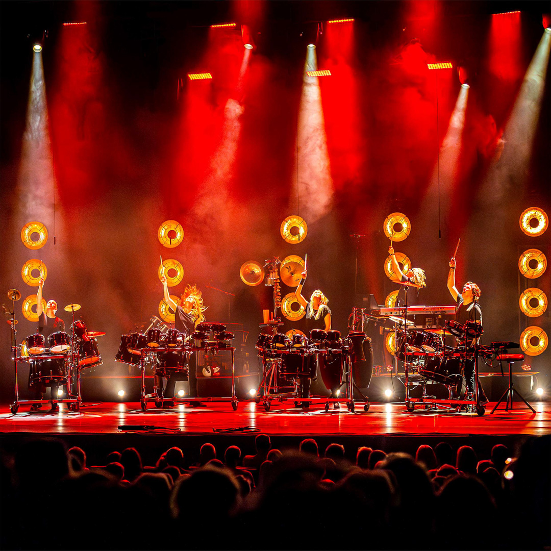 Slagwerkers van Slagerij van Kampen slaan synchroon op drums onder fel rood licht en goudkleurige spots.