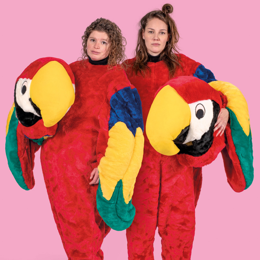 Spruijt en Opperman poseren naast elkaar in een papegaaipak met de kop onder hun arm. Zij staan voor een roze achtergrond.