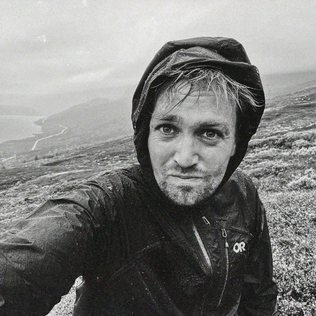 Tim Knol maakt een selfie in regenjas op een bergpad, met ruig landschap op de achtergrond.