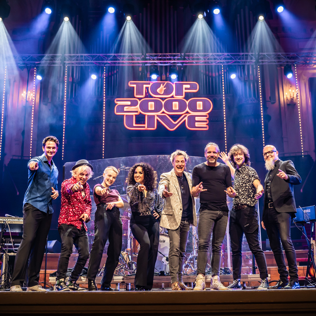 Groep artiesten poseert lachend onder fel podiumlicht met Top 2000 Live-logo achter hen.