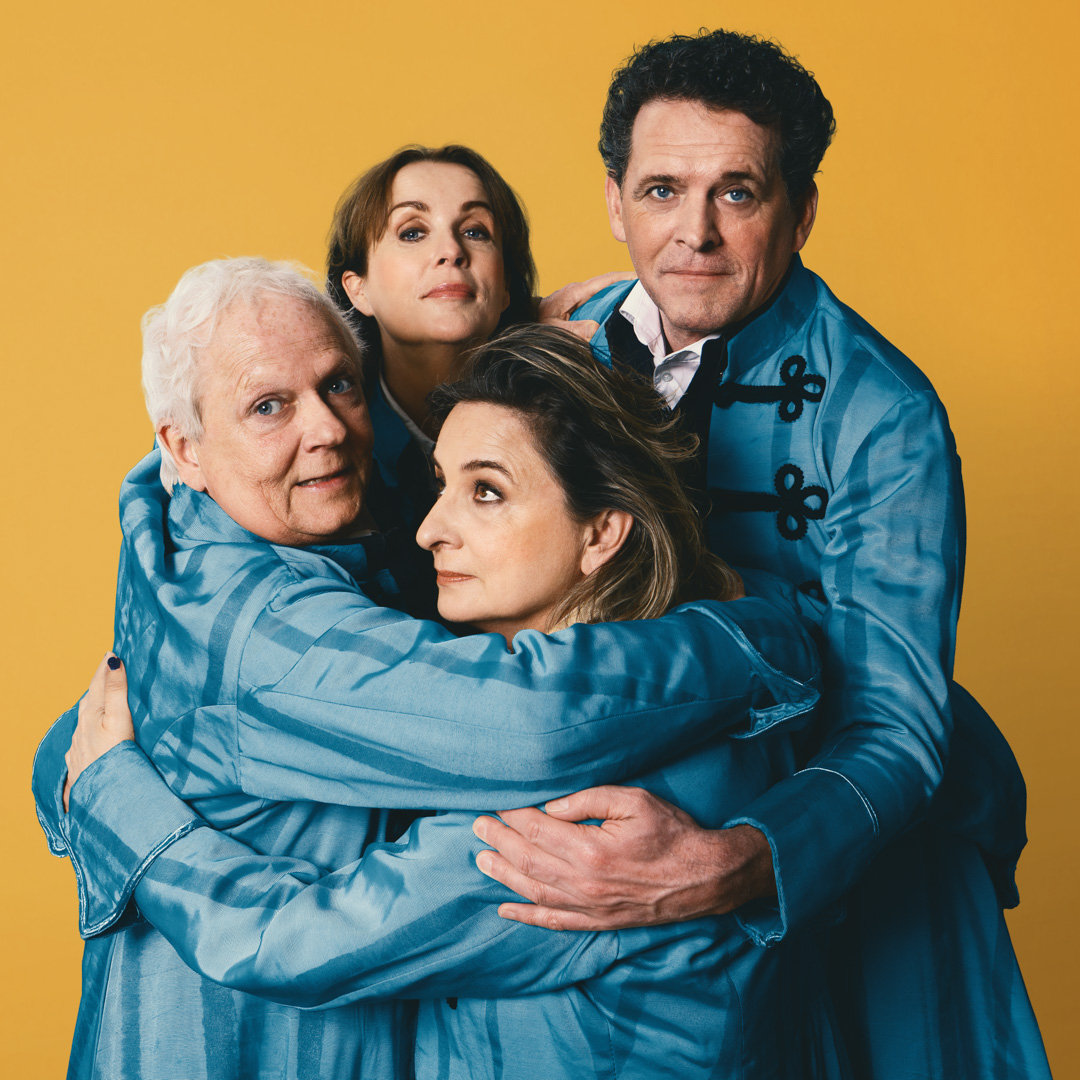 Jacqueline en Mark en twee andere acteurs in blauwe jas houden elkaar stevig vast voor een gele achtergrond.