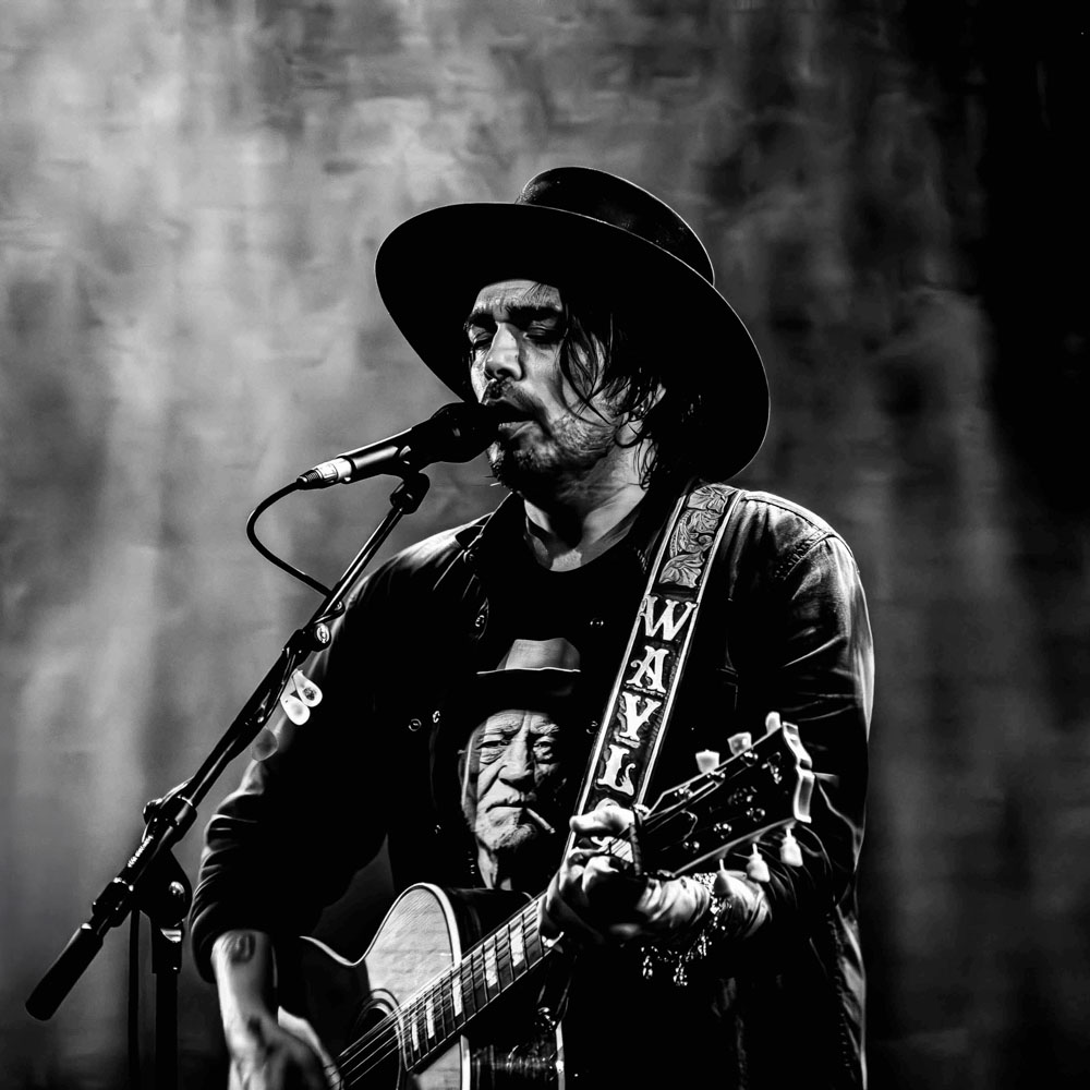 Waylon zingt met gesloten ogen en speelt gitaar in zwart-wit portret.