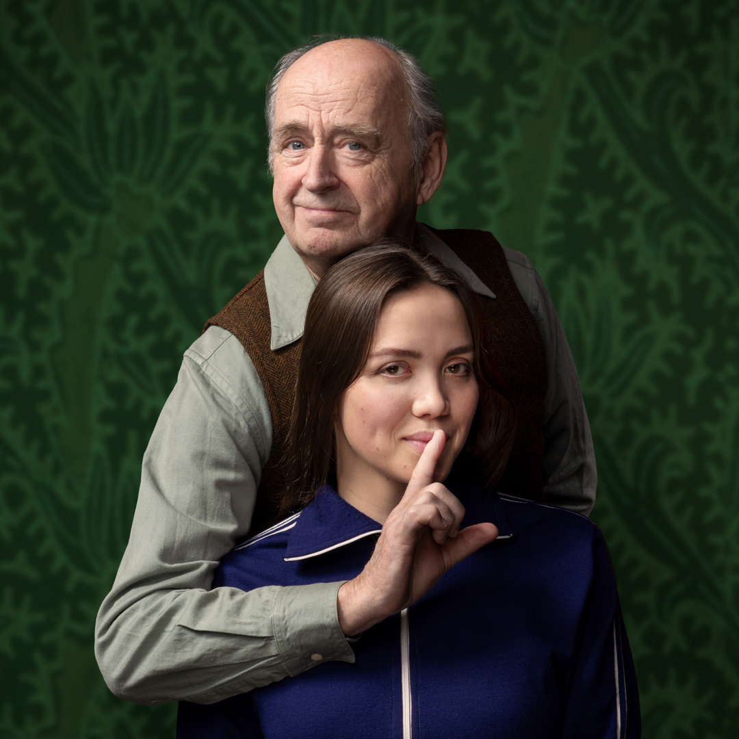 Kees Hulst en Emma Buysse poseren voor groene achtergrond, vrouw vraagt om stilte met glimlach.