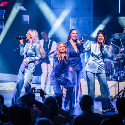 Artiesten in blauwe pakken zingen en dansen energiek voor enthousiast publiek op een fel verlicht podium.