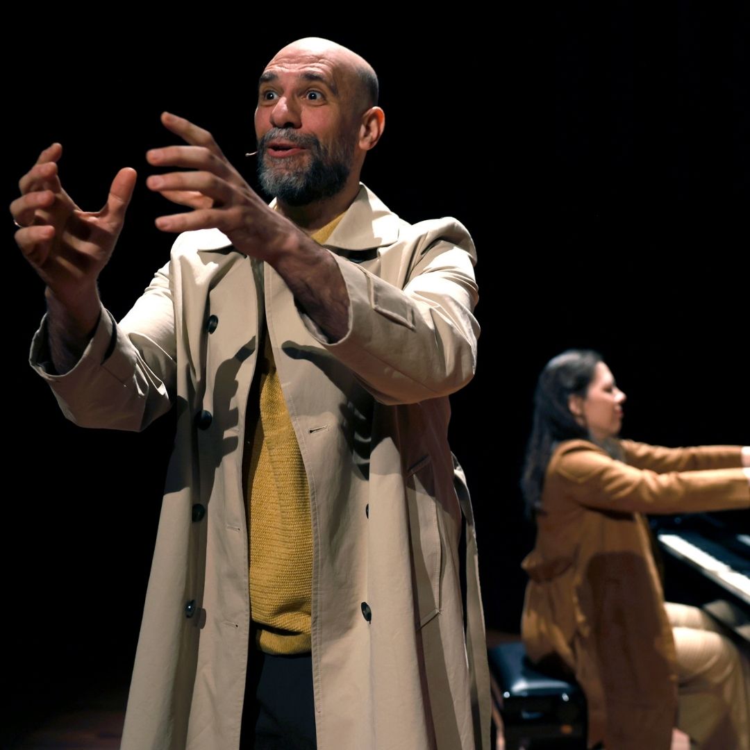 Je ziet Ali in trenchcoat gebaren in spotlicht; Nino speelt piano op de achtergrond.