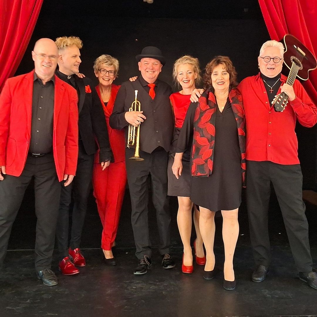 Het Nederlied Ensemble staan naast elkaar op het toneel. Ze zijn in het rood en zwart gekleed.