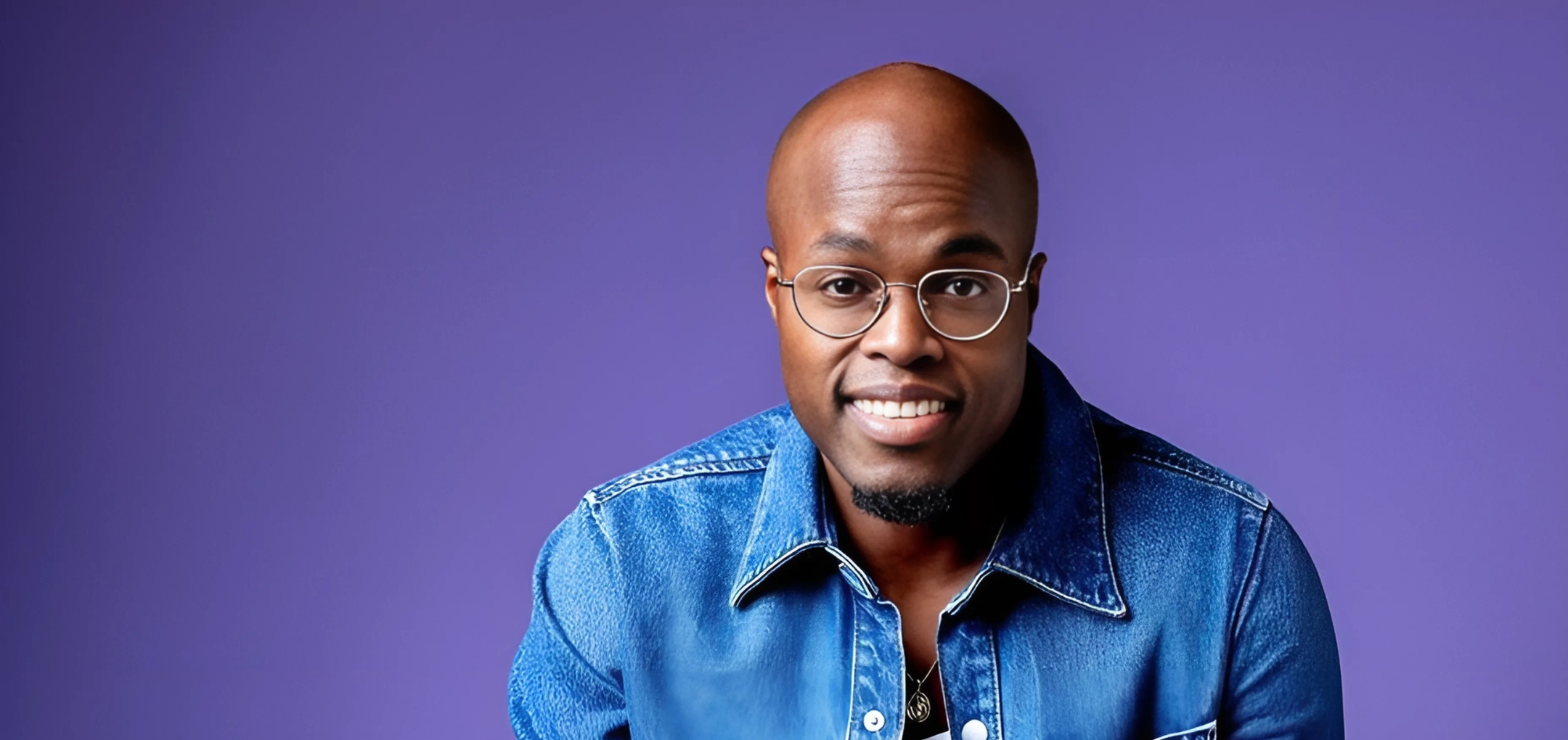 Jandino zit ontspannen op kruk in denim outfit voor paarse achtergrond.