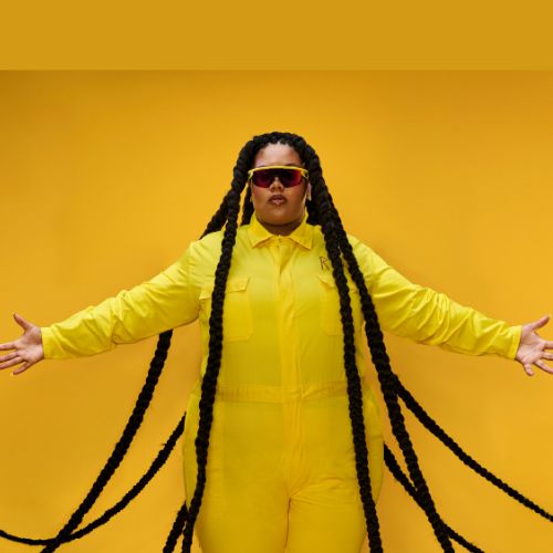 Isabelle Kafando spreid haar armen en handen uit in een gele outfit voor een gele achtergrond. Zij heeft een donkere bril op en meerdere vlechten die langs haar heen bewegen.