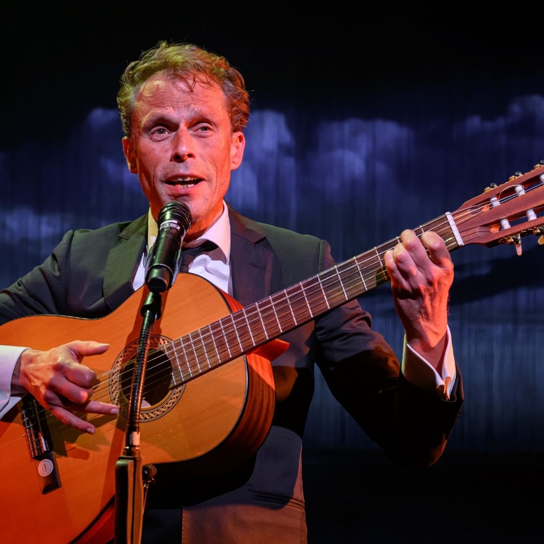 Zanger in pak speelt gitaar en zingt in de microfoon onder donkerblauw theaterlicht.