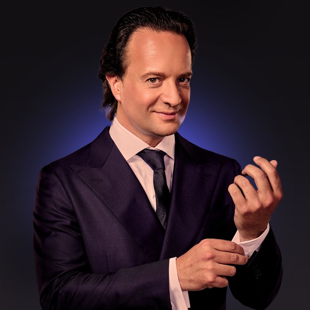 William Janz doet zijn manchetknoop goed en heeft een donkerblauwe achtergrond.