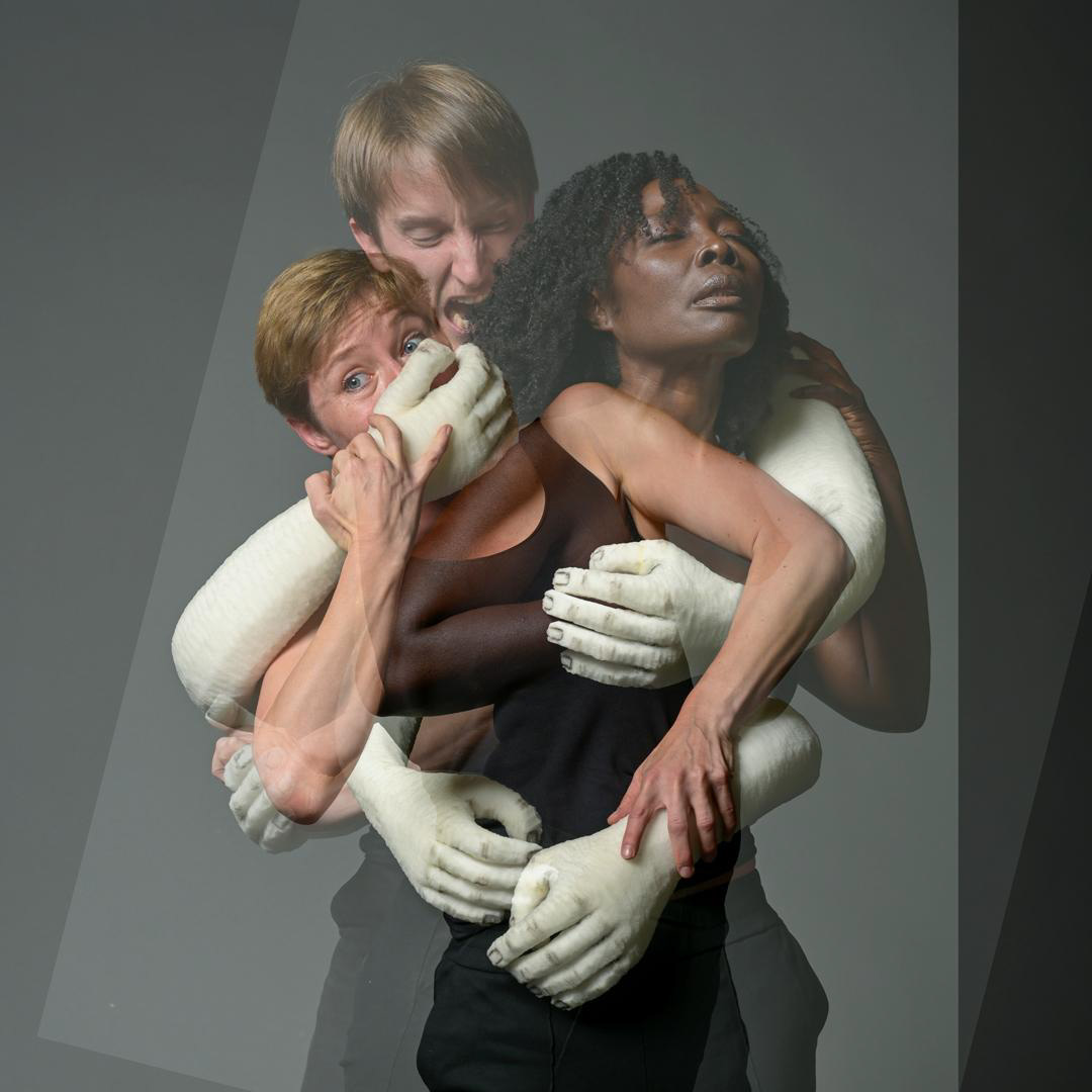 Drie performers verstrengeld, omarmd door witte poppenarmen.