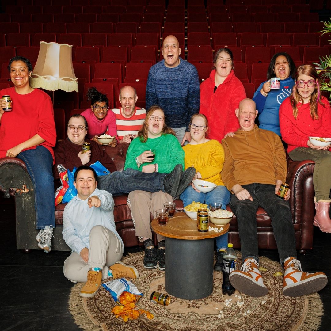 Je ziet een groep mensen in het theater zitten. De zaal is als een huiskamer aangekleed.