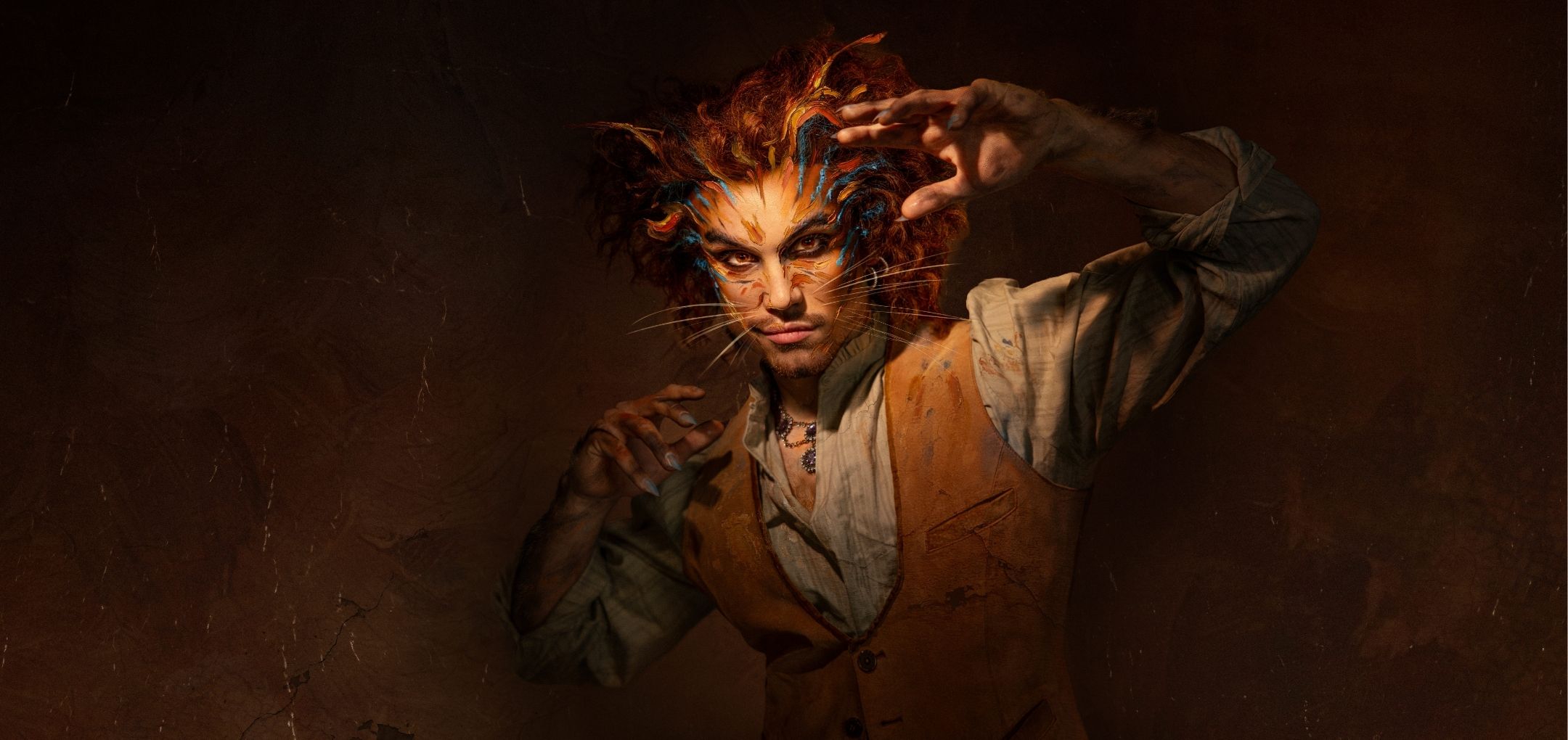 Je ziet een Cats-acteur met kattenmake-up en opgeheven handen in klauwpose onder warm licht.