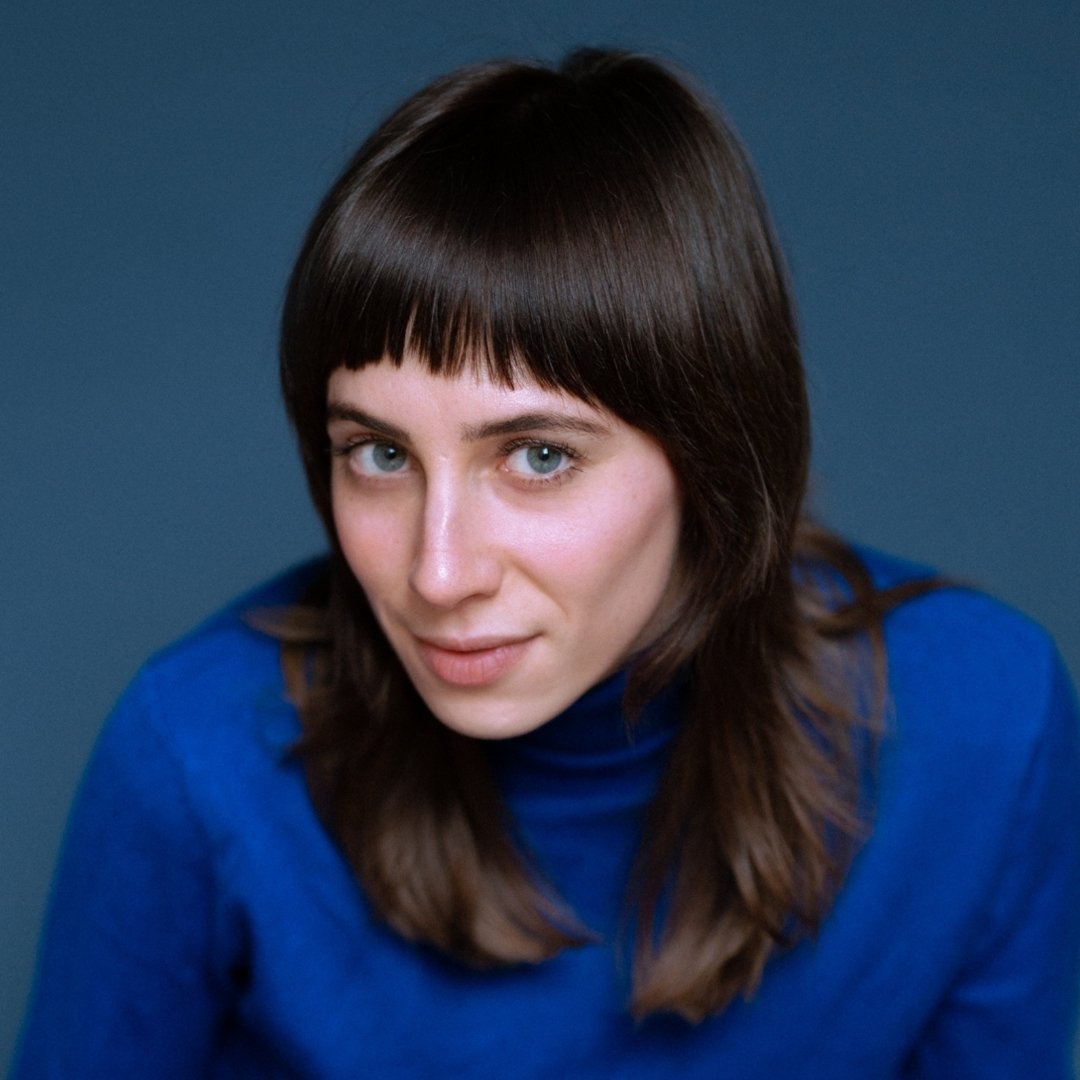 Marie Koet kijkt direct in de camera, gekleed in een blauwe coltrui, tegen een blauwe achtergrond.