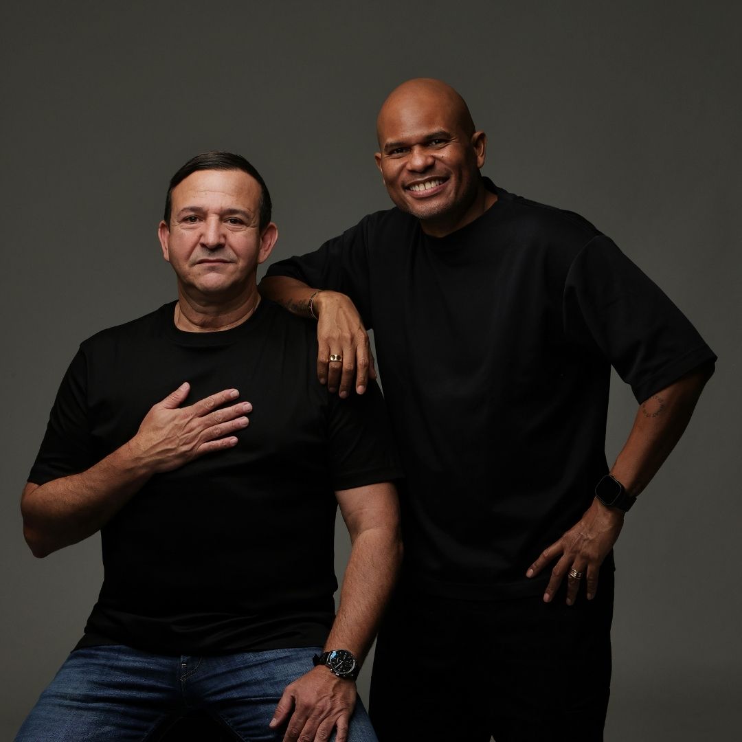 Najib Amhali zit terwijl Roue Verveer naast hem staat en lacht, beide in zwart gekleed.