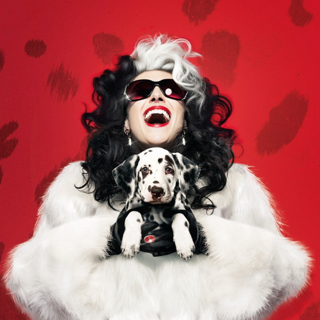Cruella staat met dalmatiër in 101 Dalmatiërs de musical tegen rode achtergrond met vlekjes.