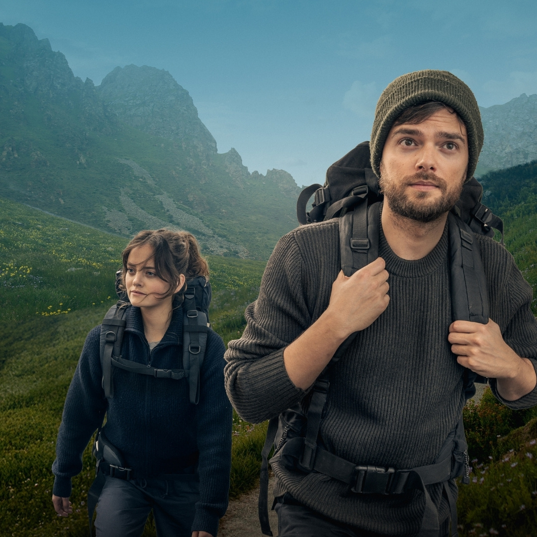 Spelers Buddy Vedder en Shelly Bos wandelen met rugzakken door een groen berglandschap.