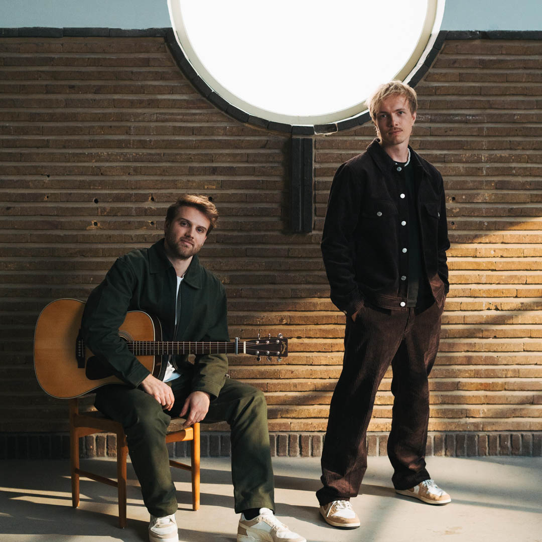 Twee muzikanten van BenR zitten met gitaar voor een rond raam in Tussen 2 maten.