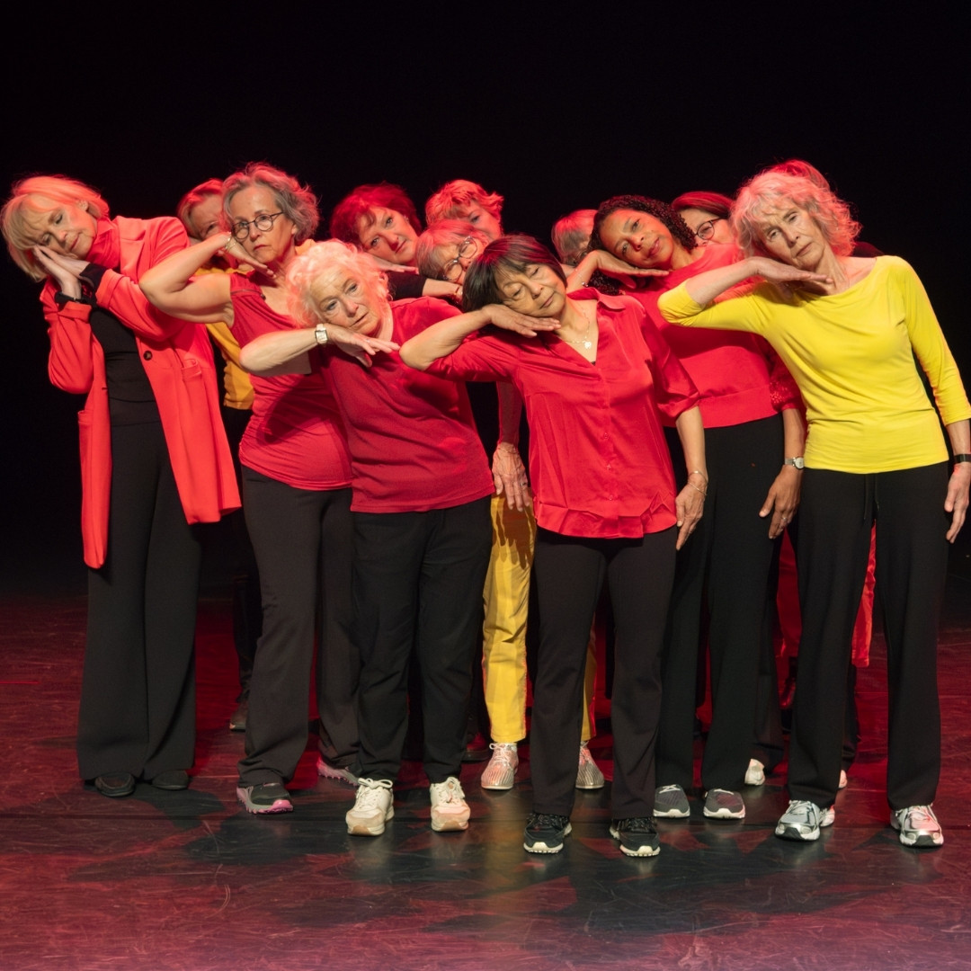 Ensemble dansers staat dicht bijeen op het podium in rode en gele kostuums, hoofd tegen schouder.