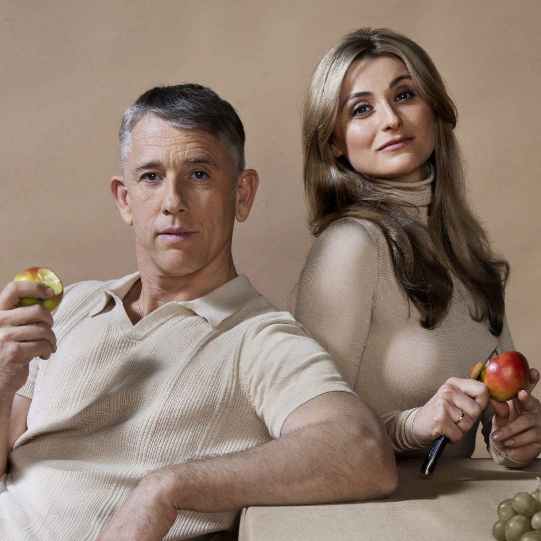 Twee spelers zitten naast elkaar in Nora met appel in de hand en indringende blik.