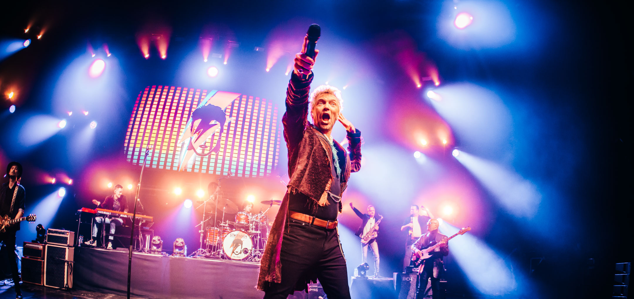 Zanger in glitterkostuum met opgeheven arm op concertpodium in Starman Bowie tribute.